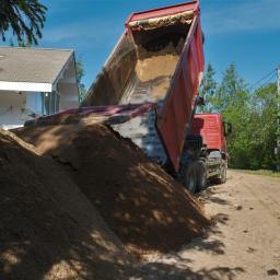 Usługi Budowlane Woźniak Jerzy - Ciężarówka z podniesioną skrzynią wyładowuje piasek na piaszczystą drogę w pobliżu domu z szarym dachem i dużymi oknami.