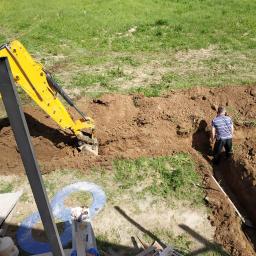 Usługi Budowlane Woźniak Jerzy - Żółta koparka wykonuje wykop liniowy w ziemi, obok stoi mężczyzna w koszulce w paski, widok z góry.