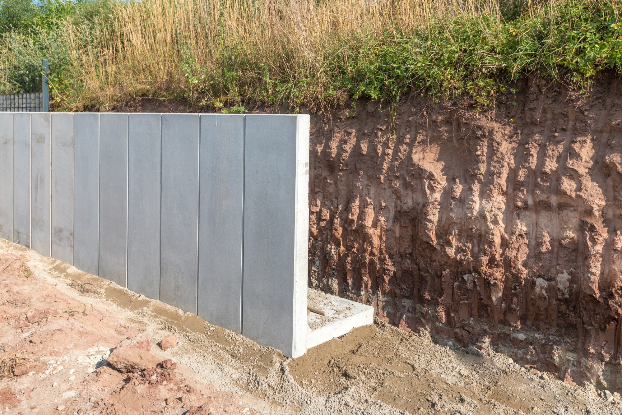 Betonowe prefabrykaty tworzące ścianę oporową w wykopie, zabezpieczające skarpę ziemną z widoczną roślinnością na górze.