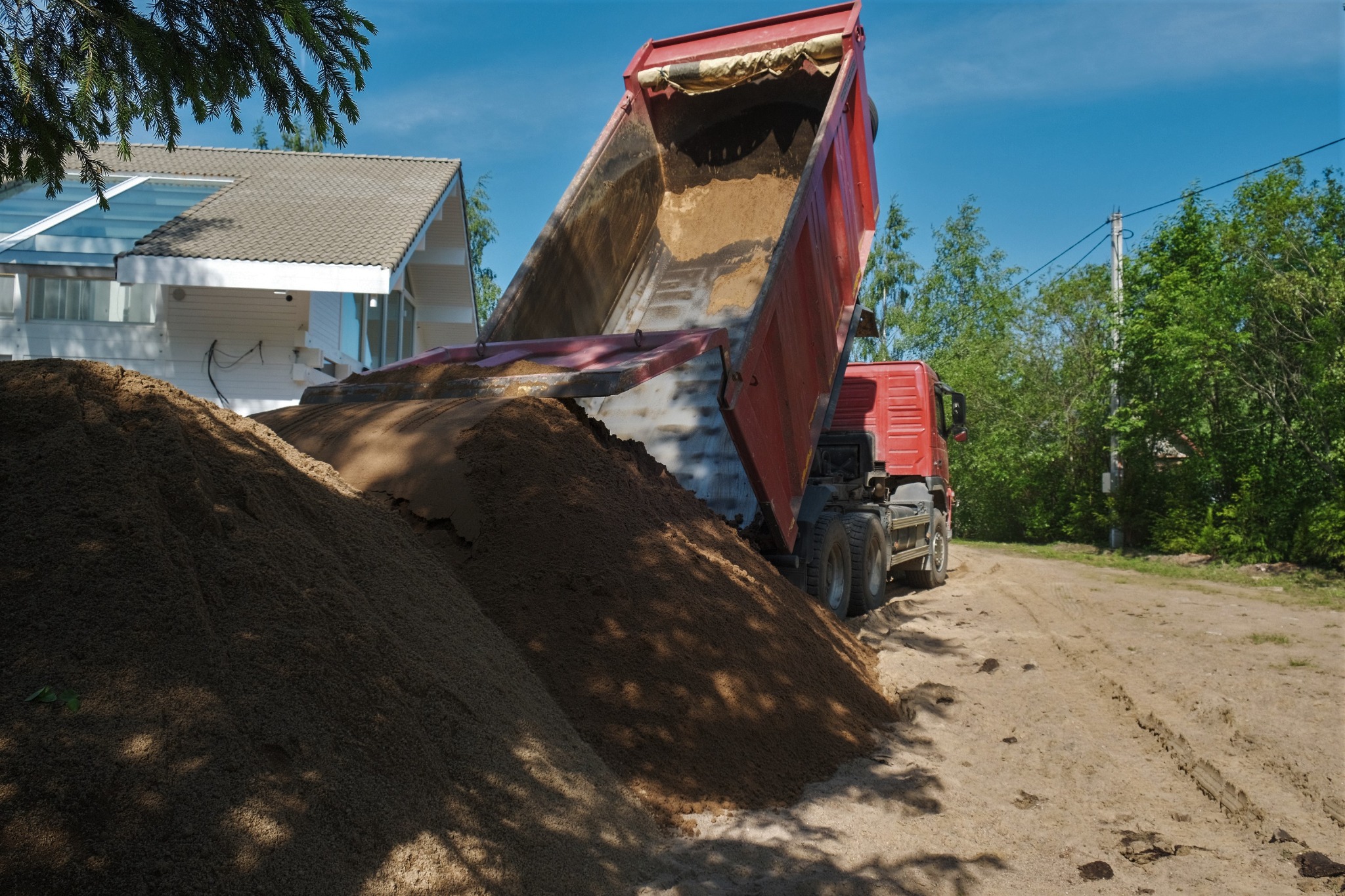 Ciężarówka z podniesioną skrzynią wyładowuje piasek na piaszczystą drogę w pobliżu domu z szarym dachem i dużymi oknami.