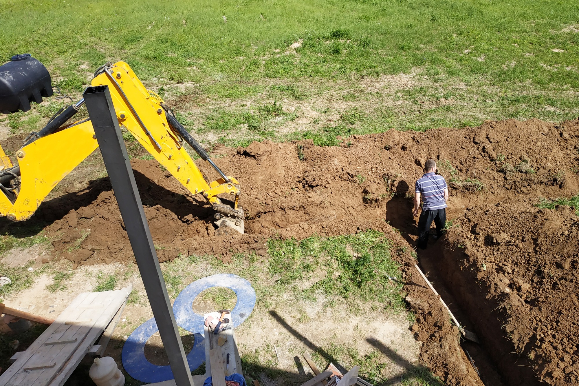 Żółta koparka wykonuje wykop liniowy w ziemi, obok stoi mężczyzna w koszulce w paski, widok z góry.