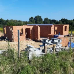 Wznoszone ściany parterowego budynku z czerwonej cegły, widoczny przenośny sanitariat i materiały budowlane na placu budowy, w tle drzewa.