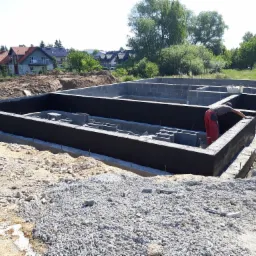 Budowa fundamentów: izolacja pionowa ścian fundamentowych z bloczków betonowych, widoczny pracownik w czerwonych spodniach, teren budowy, w tle zabudowa jednorodzinna.