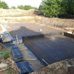 Zbrojenie fundamentu w wykopie, widoczne stalowe pręty tworzące siatkę, szalunki, narzędzia i pryzma ziemi w tle.