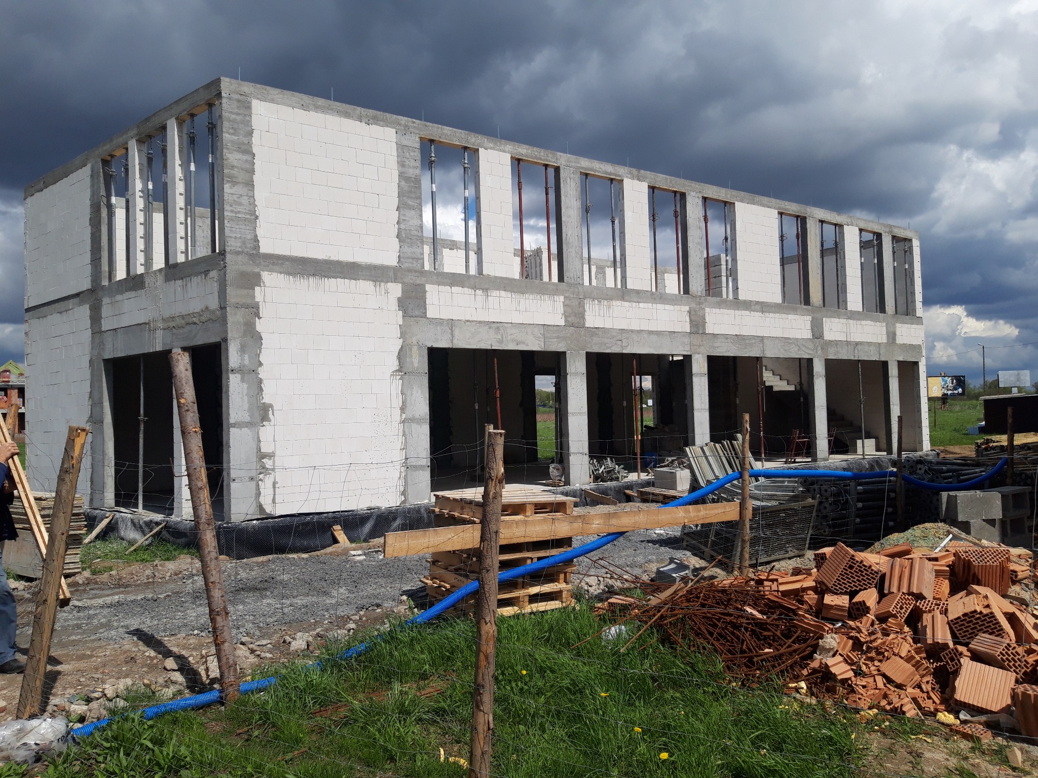 Dwukondygnacyjny budynek w stanie surowym otwartym z białych bloczków, widoczne zbrojenia i podpory, w tle burzowe chmury.