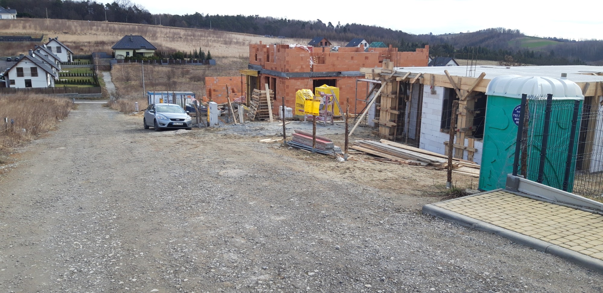 Wznoszenie ścian z czerwonej cegły na placu budowy, widoczny szalunek, przenośna toaleta i zaparkowany samochód osobowy.