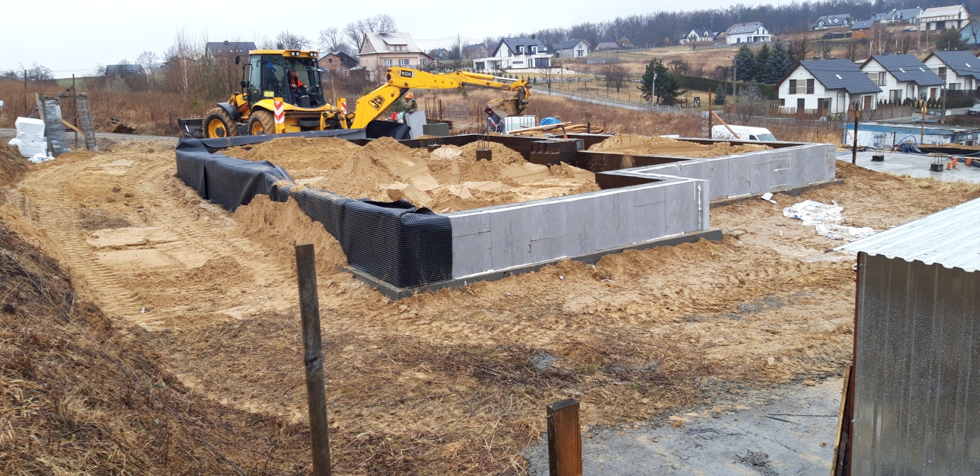 Żółta koparko-ładowarka zasypuje piaskiem fundamenty domu z szalunkiem traconym, zabezpieczone folią kubełkową, w tle zabudowa jednorodzinna.