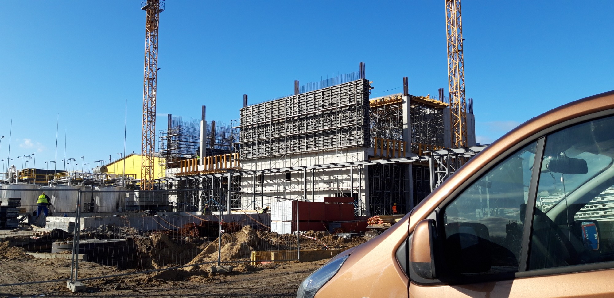 Konstrukcja budynku w trakcie budowy, widoczne szalunki, zbrojenia, żurawie, oraz fragment brązowego samochodu w pierwszym planie.