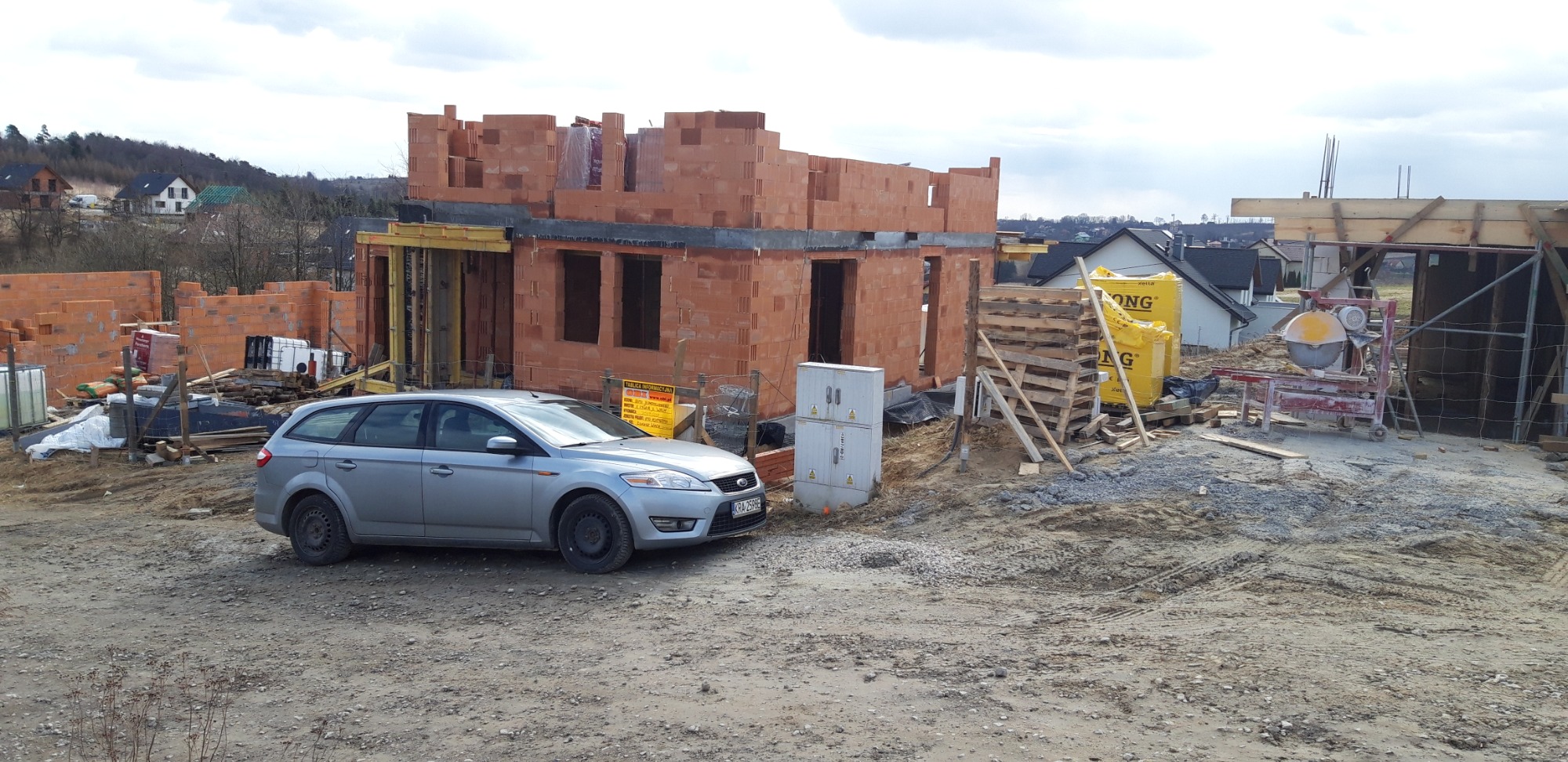 Budowa domu z cegły ceramicznej Porotherm, widoczny szary samochód osobowy Ford Focus kombi na tle placu budowy oraz częściowo wzniesione ściany budynku.