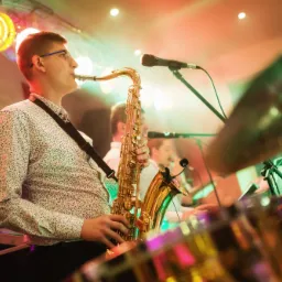 Saksofonista w koszuli we wzory gra na saksofonie na scenie, w tle widoczne perkusja i mikrofon.