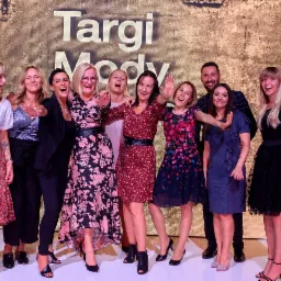 Grupa uśmiechniętych osób pozuje na tle napisu 'Targi Mody', prezentując różnorodne stylizacje odzieżowe, w tym sukienki w kwiaty, spódnice w kropki i eleganckie kombinezony.