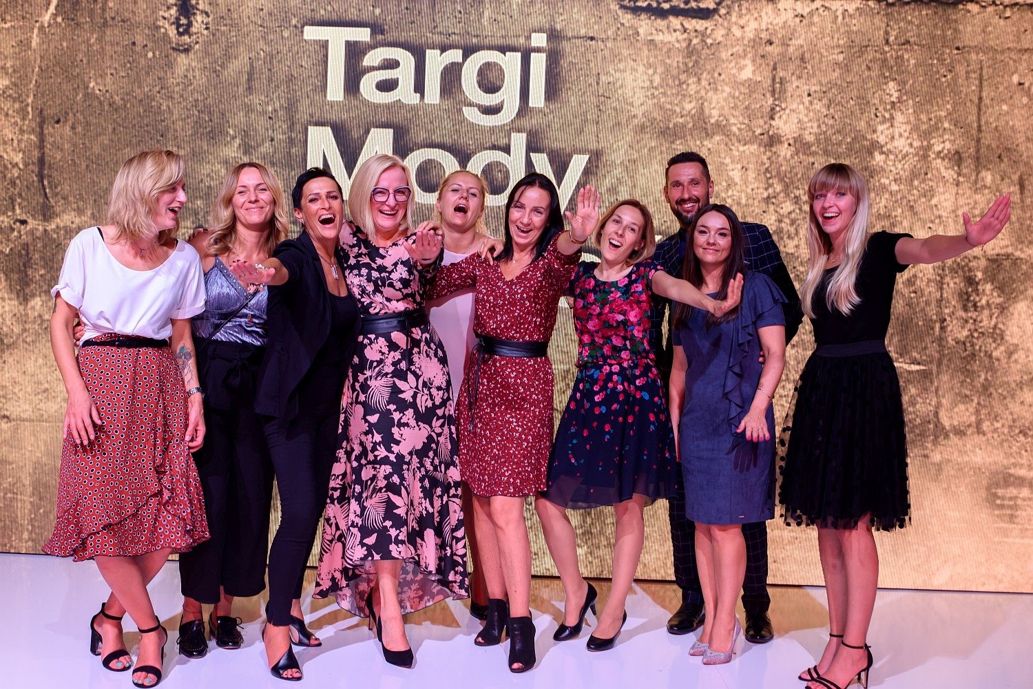 Grupa uśmiechniętych osób pozuje na tle napisu 'Targi Mody', prezentując różnorodne stylizacje odzieżowe, w tym sukienki w kwiaty, spódnice w kropki i eleganckie kombinezony.