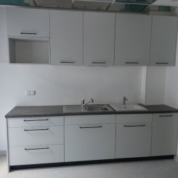 Extrmeble - Nowoczesna, jasna zabudowa kuchenna z szarym blatem i minimalistycznymi uchwytami. Widoczna umywalka i bateria. Sufit podwieszany w tle. Drzwi po prawej stronie kadru.