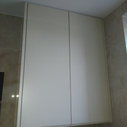 Extrmeble - Biała szafka łazienkowa z dwoma drzwiczkami, zamontowana na ścianie z beżowych płytek. Widoczny fragment sufitu i rolka papieru toaletowego.