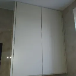 Biała szafka łazienkowa z dwoma drzwiczkami, zamontowana na ścianie z beżowych płytek. Widoczny fragment sufitu i rolka papieru toaletowego.