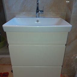 Extrmeble - Biała szafka łazienkowa z umywalką i chromowaną baterią na tle beżowych płytek. Widoczne narzędzia sugerują montaż.