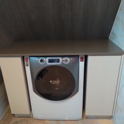 Extrmeble - Pralka Hotpoint Ariston zabudowana w szafce z blatem w łazience, widok z przodu. Jasne fronty szafek, ciemny blat i tło. Marmurowa podłoga.