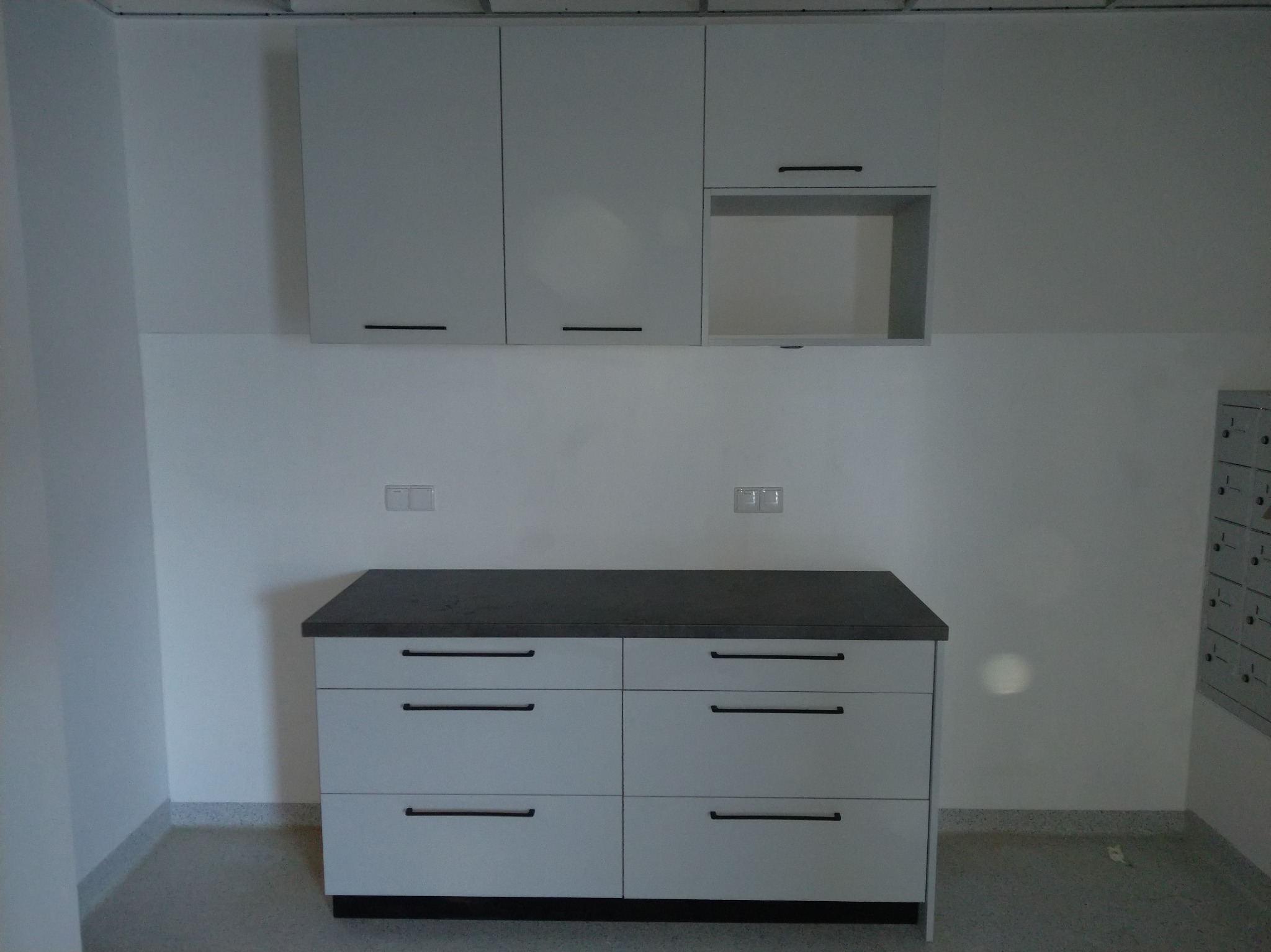 Jasnoszara zabudowa kuchenna z ciemnym blatem i czarnymi uchwytami, z szafkami wiszącymi i dolnymi szufladami, w minimalistycznym wnętrzu.