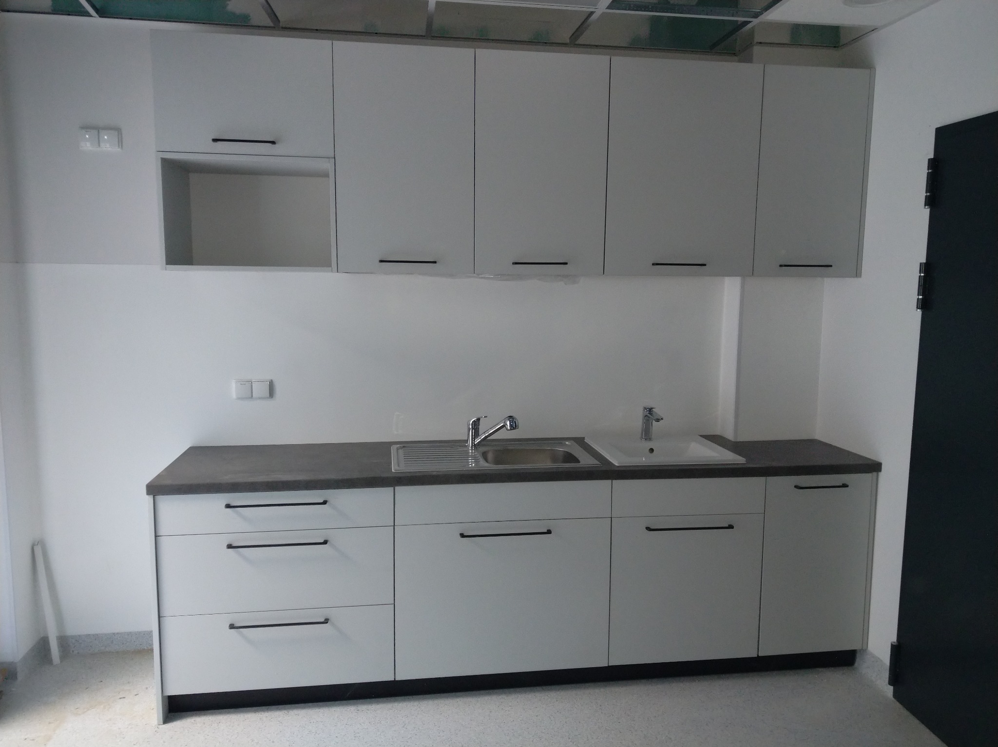 Nowoczesna, jasna zabudowa kuchenna z szarym blatem i minimalistycznymi uchwytami. Widoczna umywalka i bateria. Sufit podwieszany w tle. Drzwi po prawej stronie kadru.