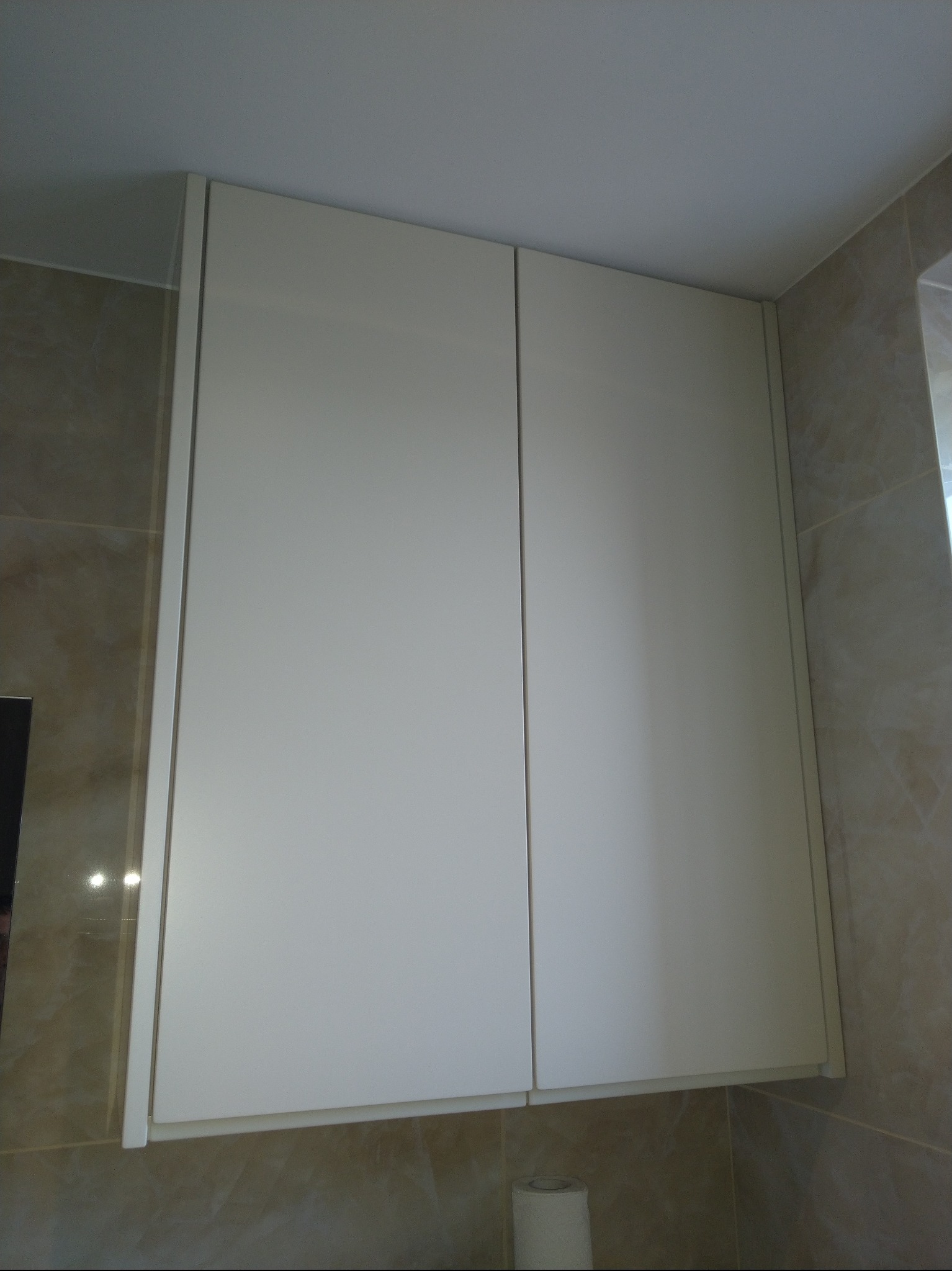 Biała szafka łazienkowa z dwoma drzwiczkami, zamontowana na ścianie z beżowych płytek. Widoczny fragment sufitu i rolka papieru toaletowego.