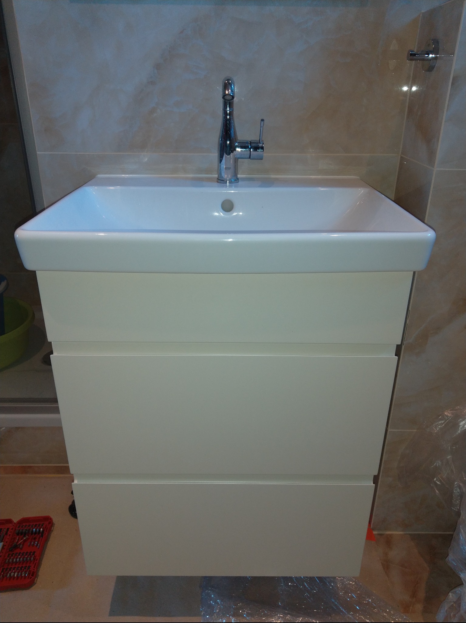 Biała szafka łazienkowa z umywalką i chromowaną baterią na tle beżowych płytek. Widoczne narzędzia sugerują montaż.