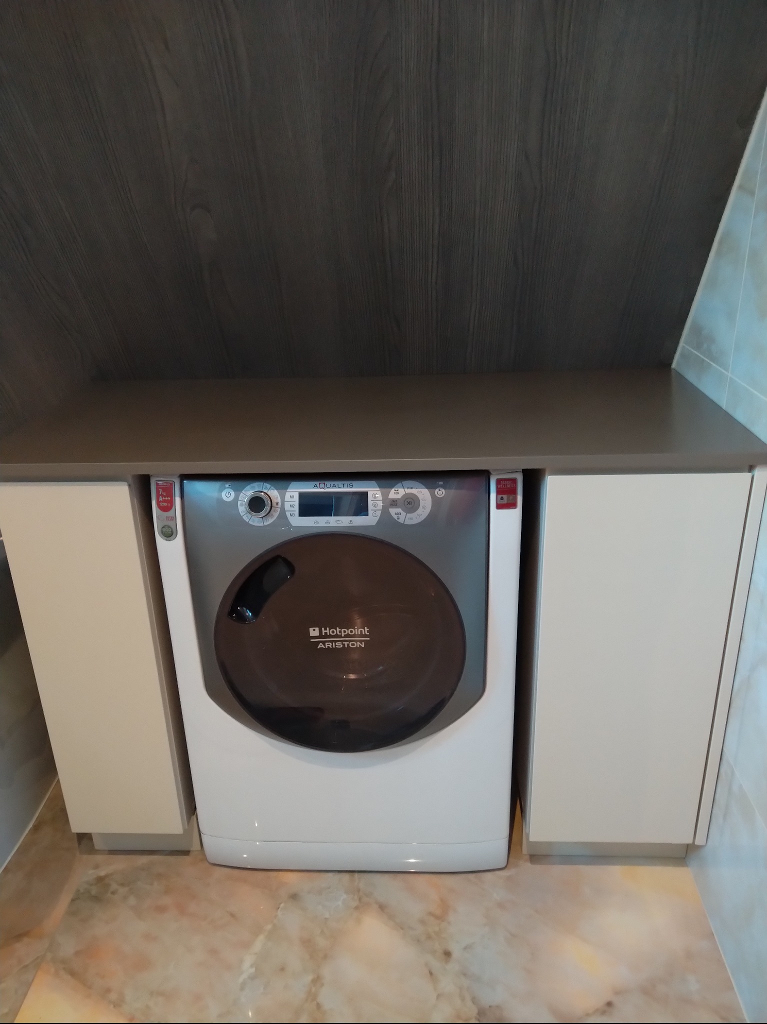 Pralka Hotpoint Ariston zabudowana w szafce z blatem w łazience, widok z przodu. Jasne fronty szafek, ciemny blat i tło. Marmurowa podłoga.