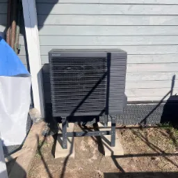 Zewnętrzna jednostka pompy ciepła Panasonic Aquarea zamontowana na betonowych podstawach przy szarej elewacji budynku. Widoczne elementy instalacji i cień.