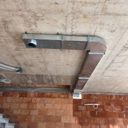 Frigus Instalacje - Kanciasta, metalowa instalacja wentylacyjna przymocowana do sufitu w surowym wnętrzu z czerwonej cegły. Widoczny fragment schodów i rusztowania.