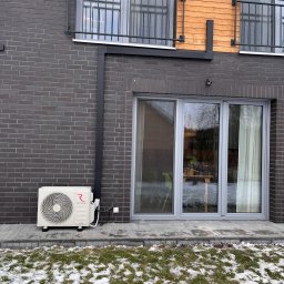Frigus Instalacje - Biała jednostka zewnętrzna klimatyzacji Rotenso Hiro zamontowana na elewacji z ciemnej cegły, widoczna trawa z resztkami śniegu i okno tarasowe.