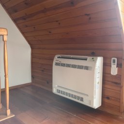 Frigus Instalacje - Biała klimatyzacja podłogowa Gree z pilotem na drewnianej ścianie i podłodze w pokoju na poddaszu. Widoczny fragment schodów.