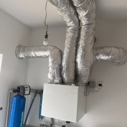 Frigus Instalacje - Instalacja rekuperacji z widocznymi srebrnymi rurami wentylacyjnymi pod sufitem i białym centralnym urządzeniem. Dodatkowo widoczny filtr wody.