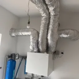 Instalacja rekuperacji z widocznymi srebrnymi rurami wentylacyjnymi pod sufitem i białym centralnym urządzeniem. Dodatkowo widoczny filtr wody.