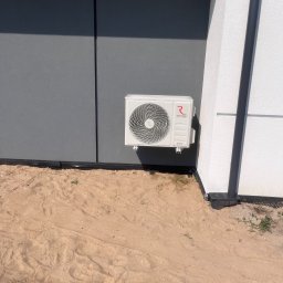 Frigus Instalacje - Biała jednostka zewnętrzna klimatyzacji Rotenso zamontowana na szarej ścianie budynku, z widocznym odpływem skroplin i piaskowym podłożem.