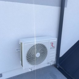 Frigus Instalacje - Biała jednostka zewnętrzna klimatyzacji Rotenso Teta Series zamontowana na ścianie budynku, z widoczną rurą spustową i fragmentem dachu w tle.