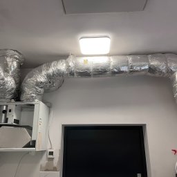 Frigus Instalacje - Kompletny system wentylacyjny z srebrnymi rurami zamontowany pod sufitem, obok biała jednostka centralna i panel sterowania na ścianie.