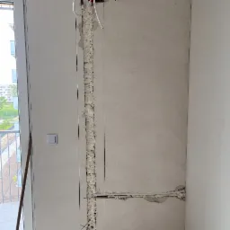 Wnętrze w trakcie prac: wycięty pionowy rowek w ścianie wypełniony pianką montażową, widoczne przewody i rurki. Fragment okna balkonowego z widokiem na zewnątrz.