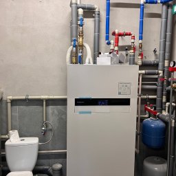 Frigus Instalacje - Biała pompa ciepła Panasonic Aquarea z licznymi rurami (szare, niebieskie, czerwone) w pomieszczeniu technicznym. Widoczny fragment toalety. Szare ściany.