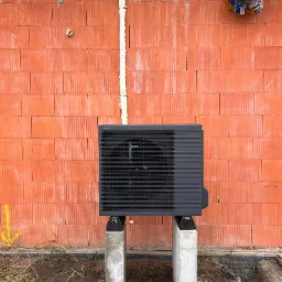 Czarna pompa ciepła Panasonic zamontowana na betonowych wspornikach przed ceglaną ścianą budynku w trakcie budowy. Widoczne rury i izolacja.