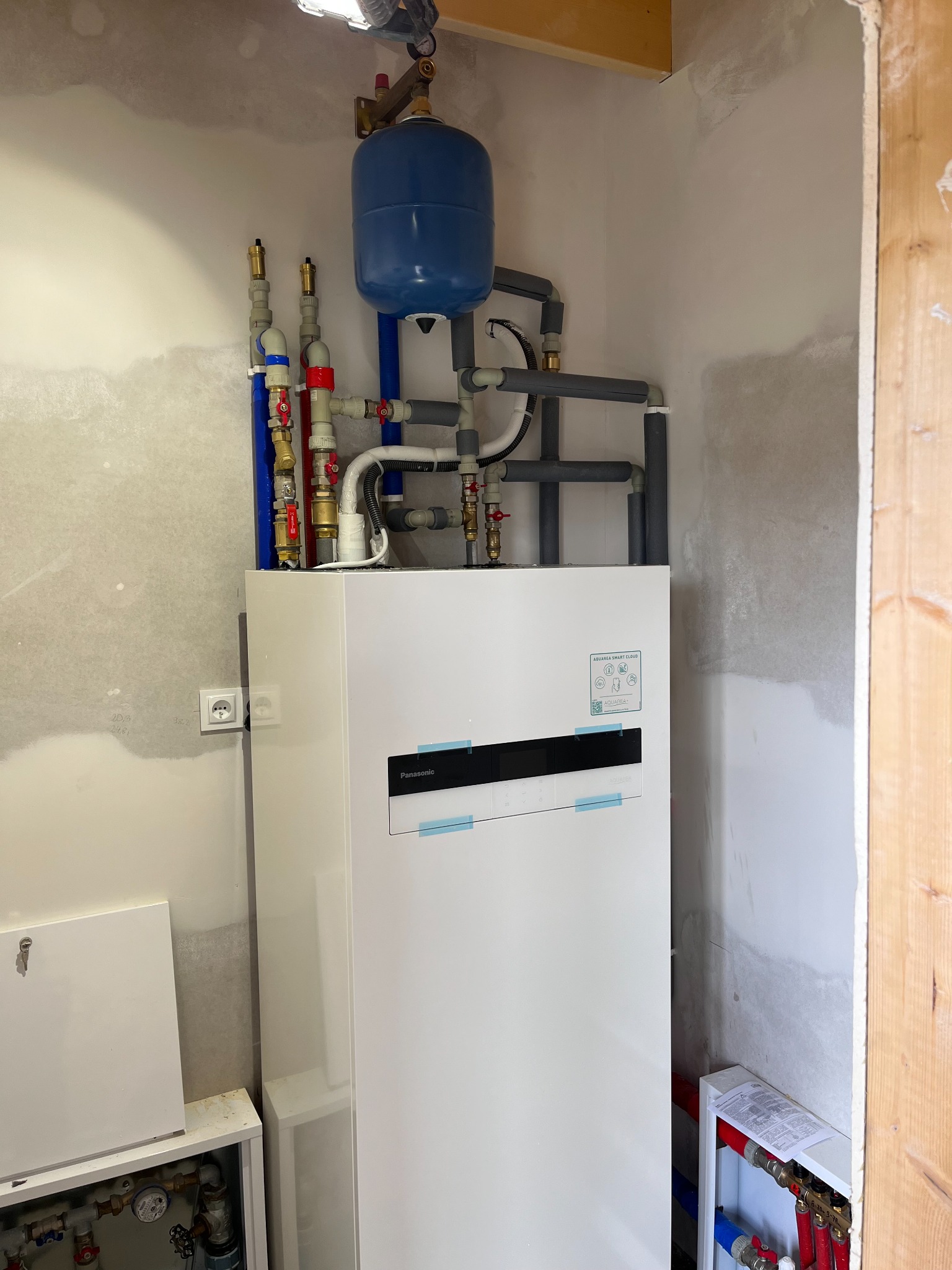 Biała pompa ciepła Panasonic z niebieskim zbiornikiem wyrównawczym i instalacją hydrauliczną w surowym wnętrzu. Widoczny fragment ściany z gładzią i drewniane wykończenie.