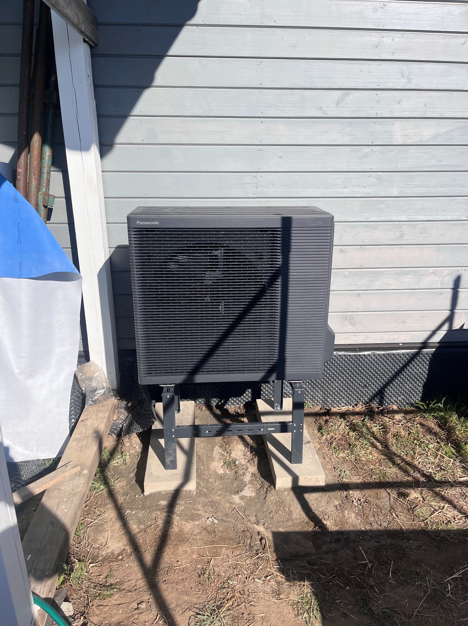 Zewnętrzna jednostka pompy ciepła Panasonic Aquarea zamontowana na betonowych podstawach przy szarej elewacji budynku. Widoczne elementy instalacji i cień.