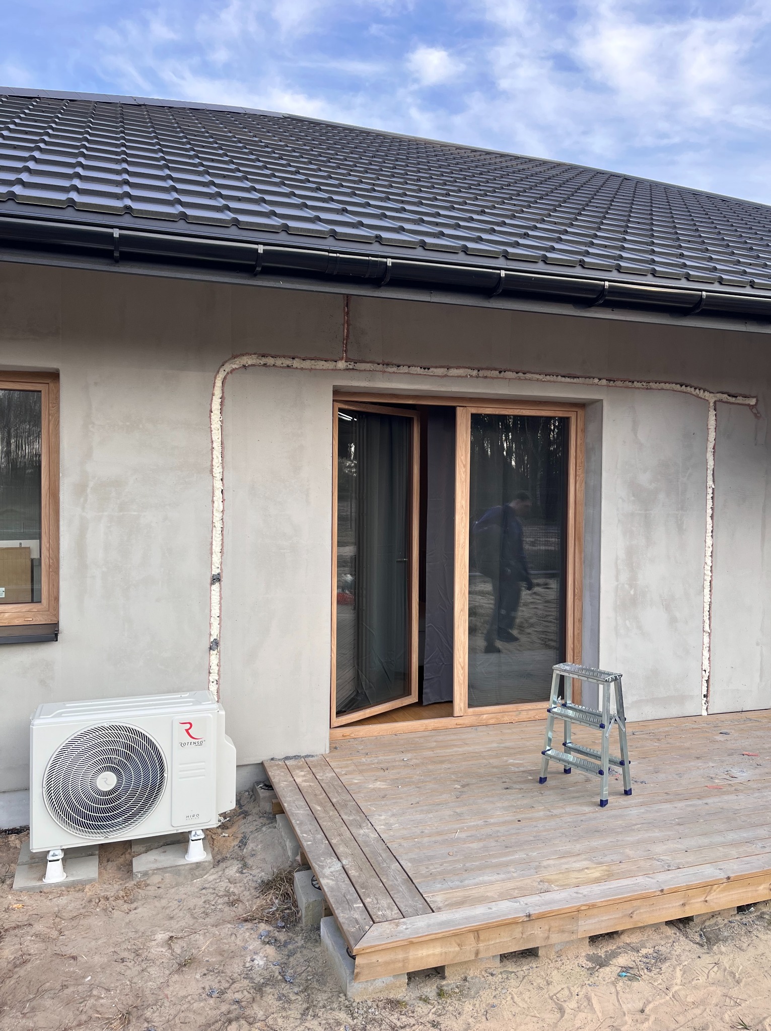 Montaż klimatyzacji Rotenso na elewacji domu z drewnianą stolarką okienną i zarysowanymi liniami pod instalacje. Widoczny fragment drewnianego tarasu i drabina.