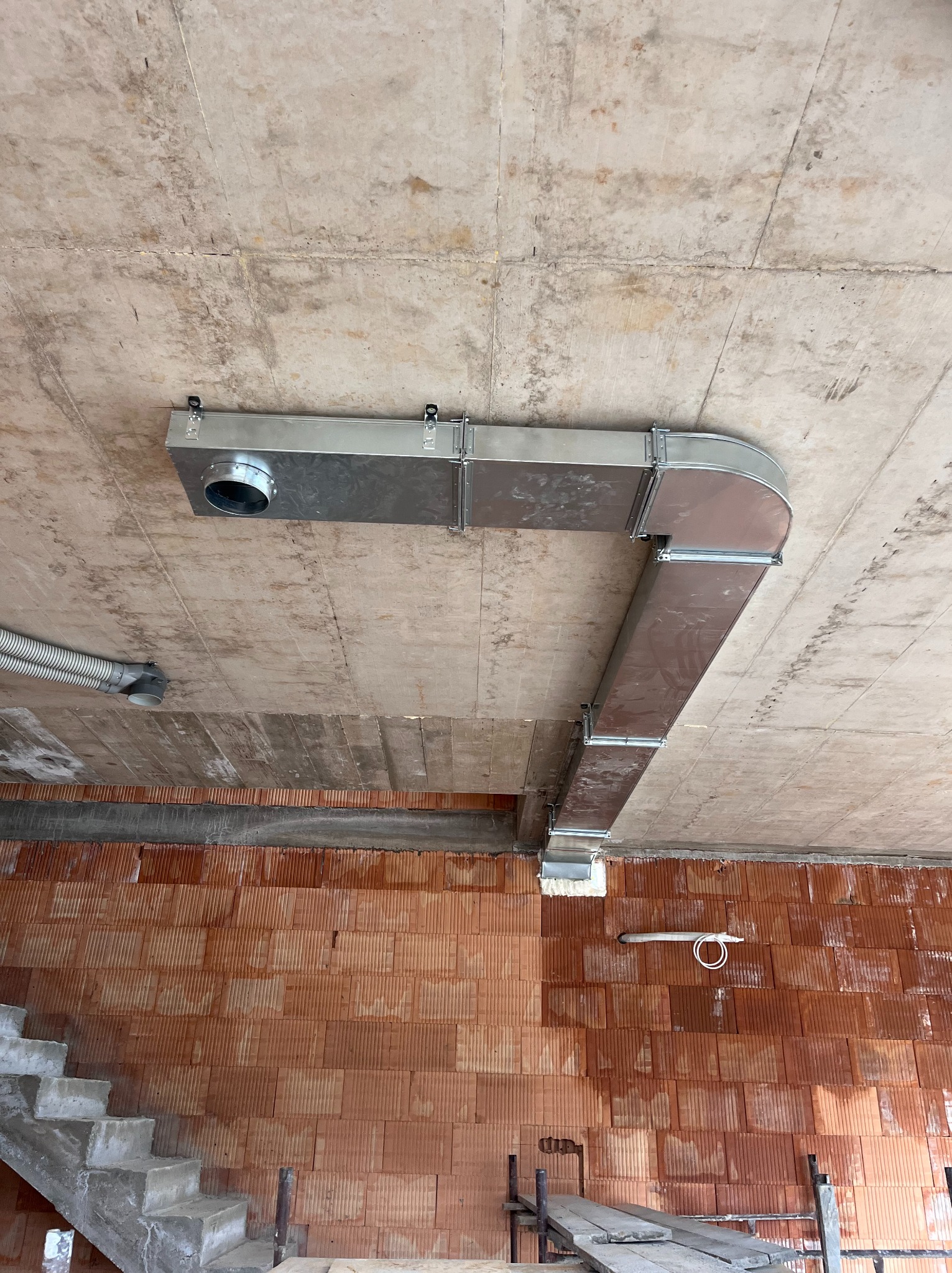 Kanciasta, metalowa instalacja wentylacyjna przymocowana do sufitu w surowym wnętrzu z czerwonej cegły. Widoczny fragment schodów i rusztowania.