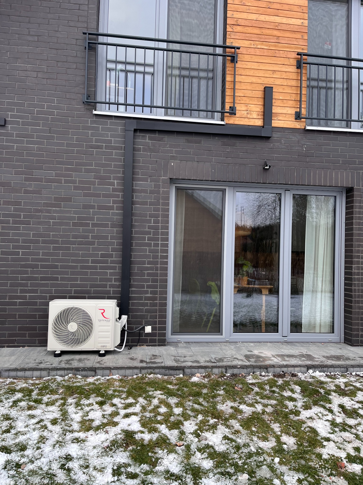 Biała jednostka zewnętrzna klimatyzacji Rotenso Hiro zamontowana na elewacji z ciemnej cegły, widoczna trawa z resztkami śniegu i okno tarasowe.