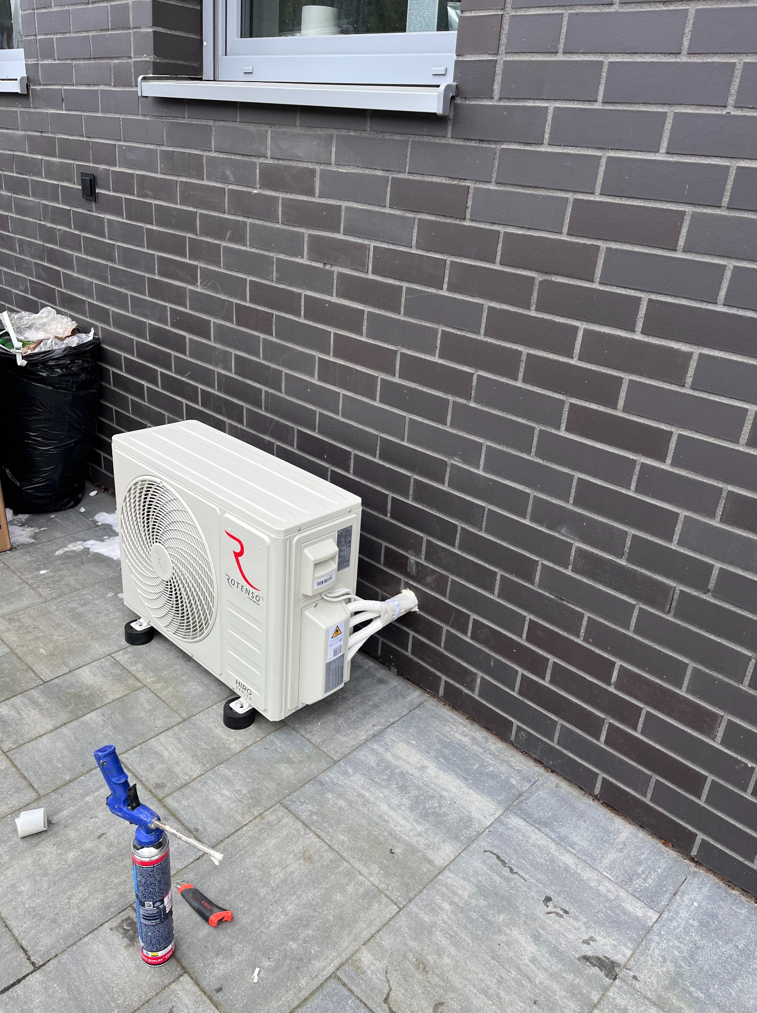 Biała jednostka zewnętrzna klimatyzacji Rotenso, zamontowana przy ceglanej ścianie budynku, z widocznymi narzędziami montażowymi na szarej kostce brukowej.