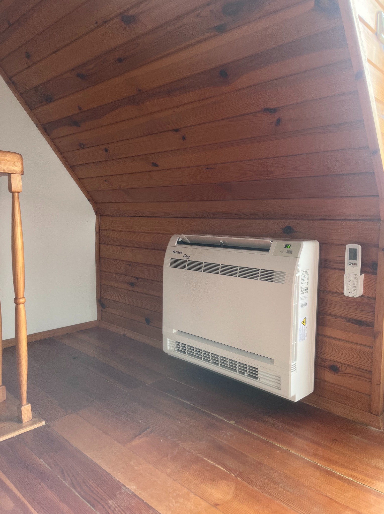 Biała klimatyzacja podłogowa Gree z pilotem na drewnianej ścianie i podłodze w pokoju na poddaszu. Widoczny fragment schodów.