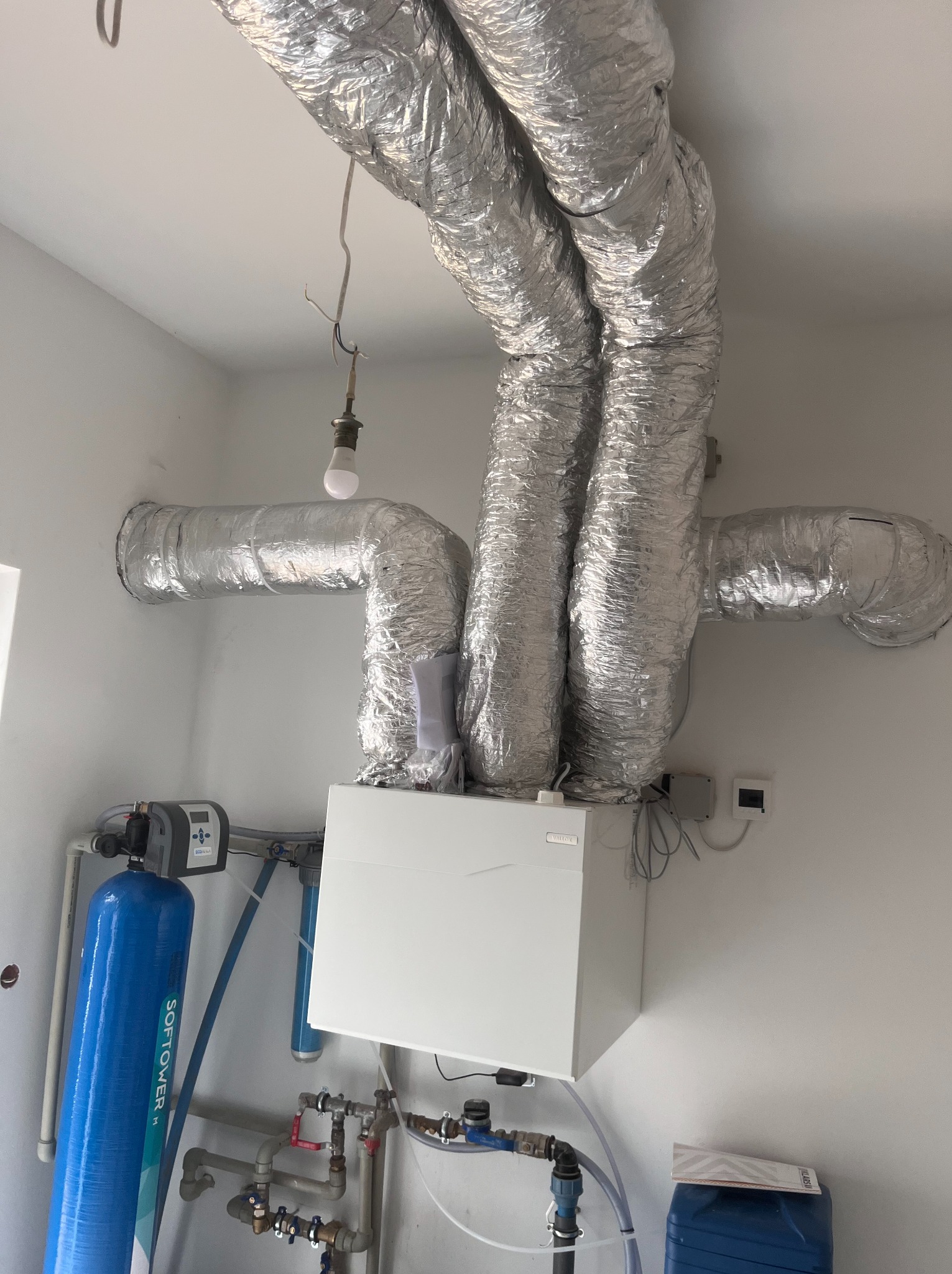 Instalacja rekuperacji z widocznymi srebrnymi rurami wentylacyjnymi pod sufitem i białym centralnym urządzeniem. Dodatkowo widoczny filtr wody.