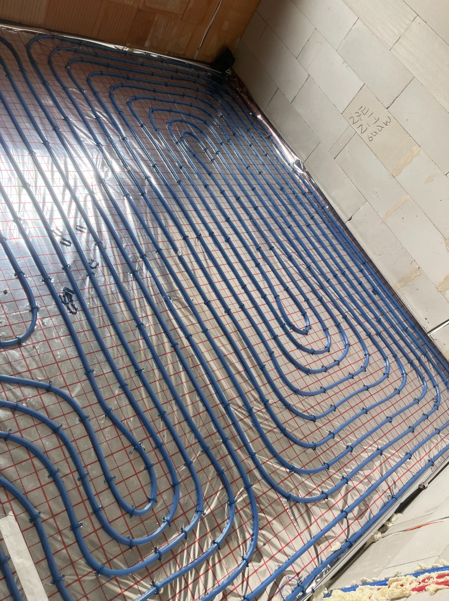 Niebieskie rury ogrzewania podłogowego ułożone na folii z siatką, zamocowane plastikowymi klipsami. Widok z góry, surowy stan, ściany z bloków.