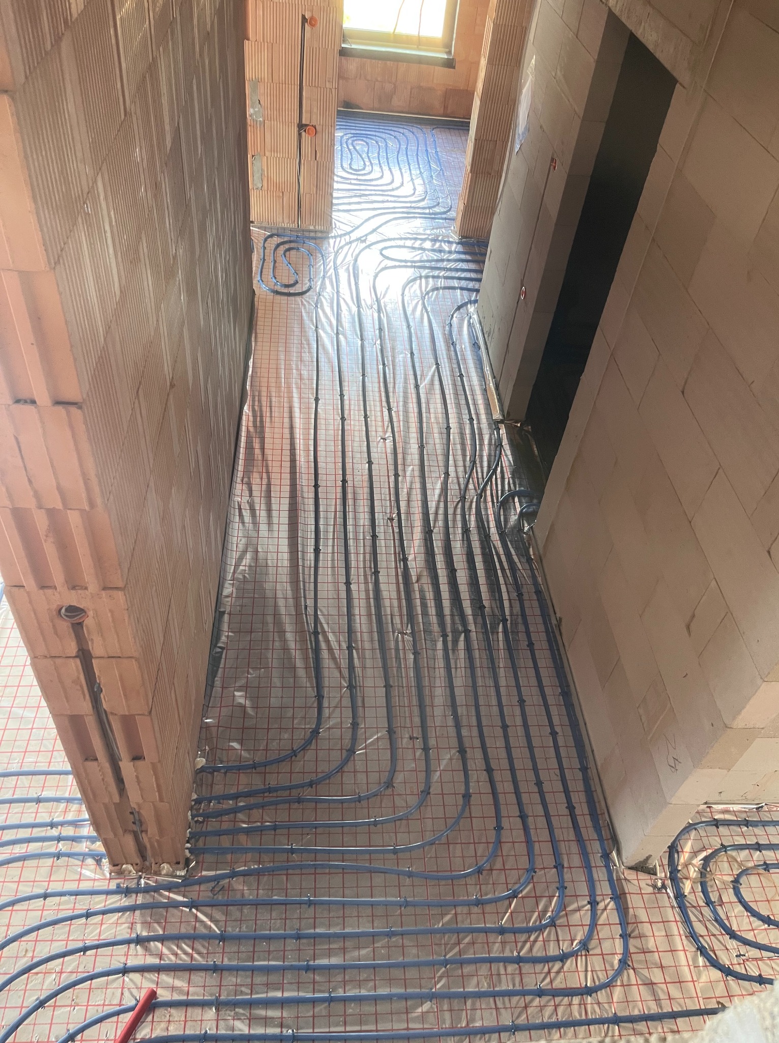 Niebieskie rurki ogrzewania podłogowego ułożone na folii z siatką w surowym wnętrzu budynku z ceglanymi ścianami, widok z góry.