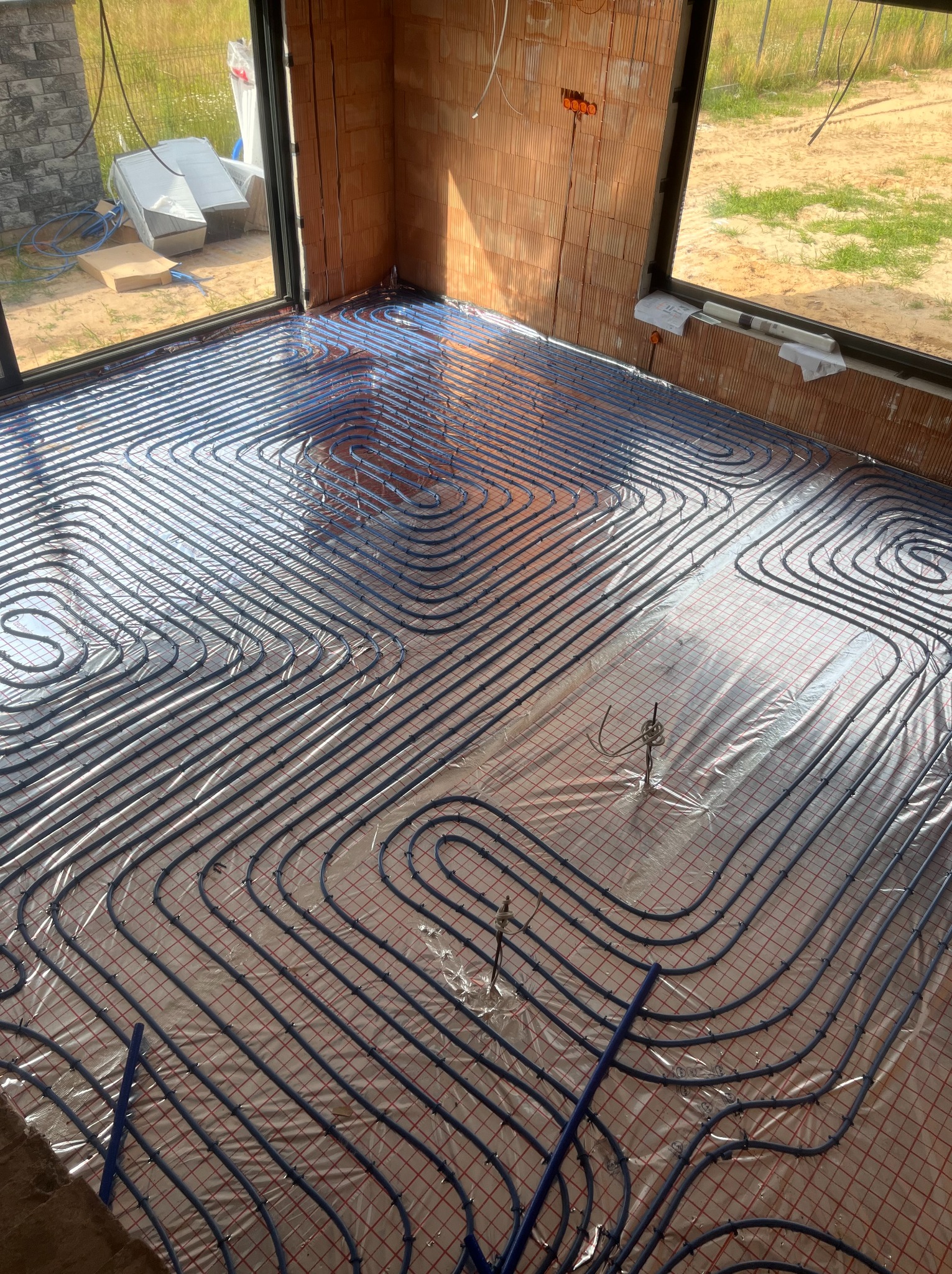 Instalacja ogrzewania podłogowego w surowym wnętrzu z ceglanymi ścianami. Równomiernie rozłożone rury grzewcze układają się w regularny wzór na folii izolacyjnej.