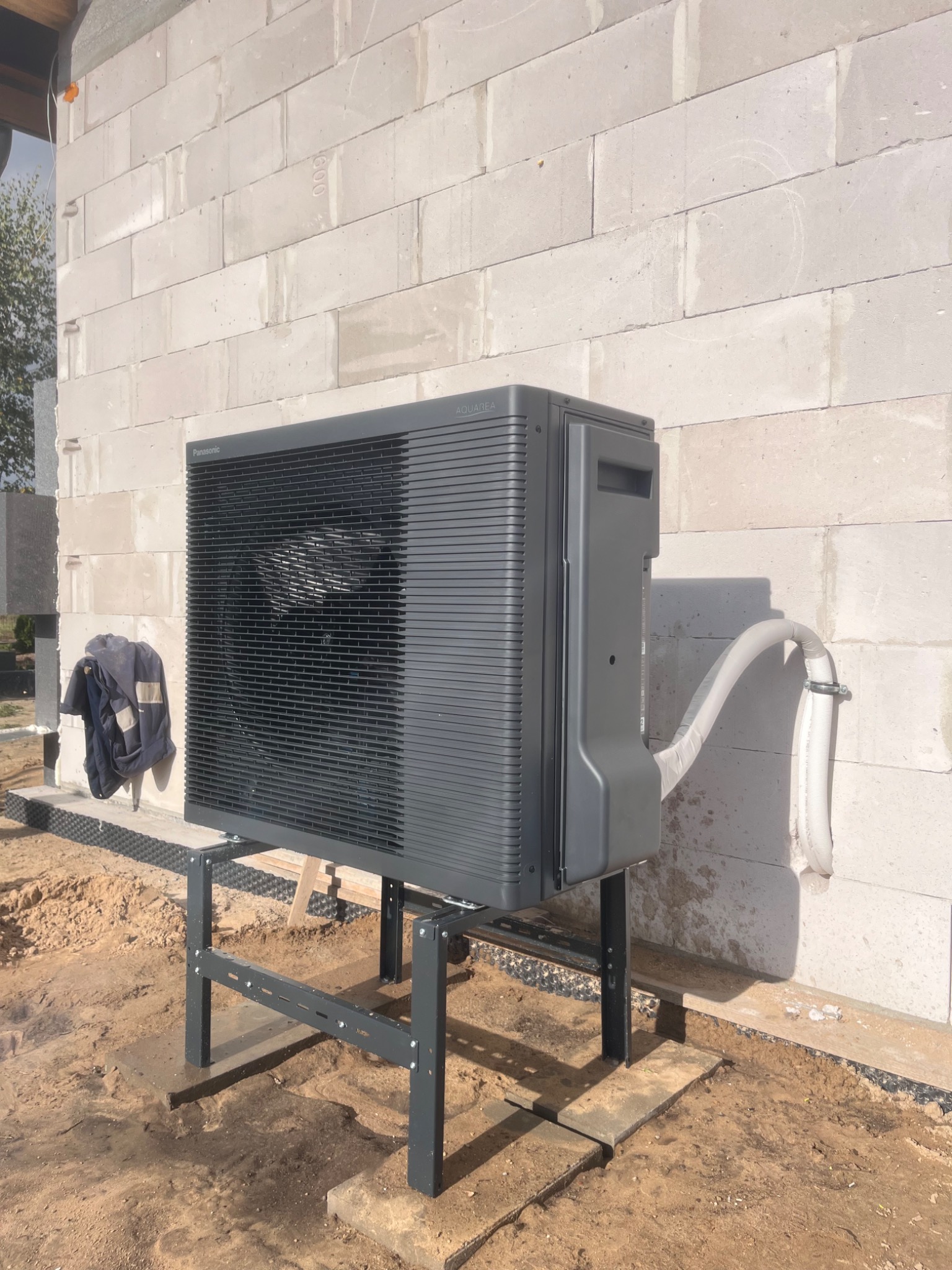 Szara pompa ciepła Panasonic Aquarea zamontowana na metalowym stelażu przy ścianie z betonu komórkowego. Widoczne przyłącza i robocza odzież.